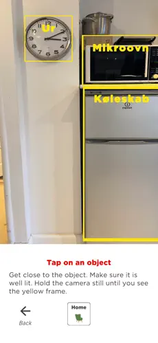 A screenshot from the Speakit app, showing the main camera UI with three objects highlighted and labelled in Danish: Ur, Mikroovn and Køleskab. Underneath is the text "Tap on an object. Get close to the object. Make sure it is well lit.Hold the camera still until you see the yellow frame." Below are two buttons, from left to right: a button with a left-facing arrow and the label "Back", an outlined button with an armchair silhouette and the label "Home".